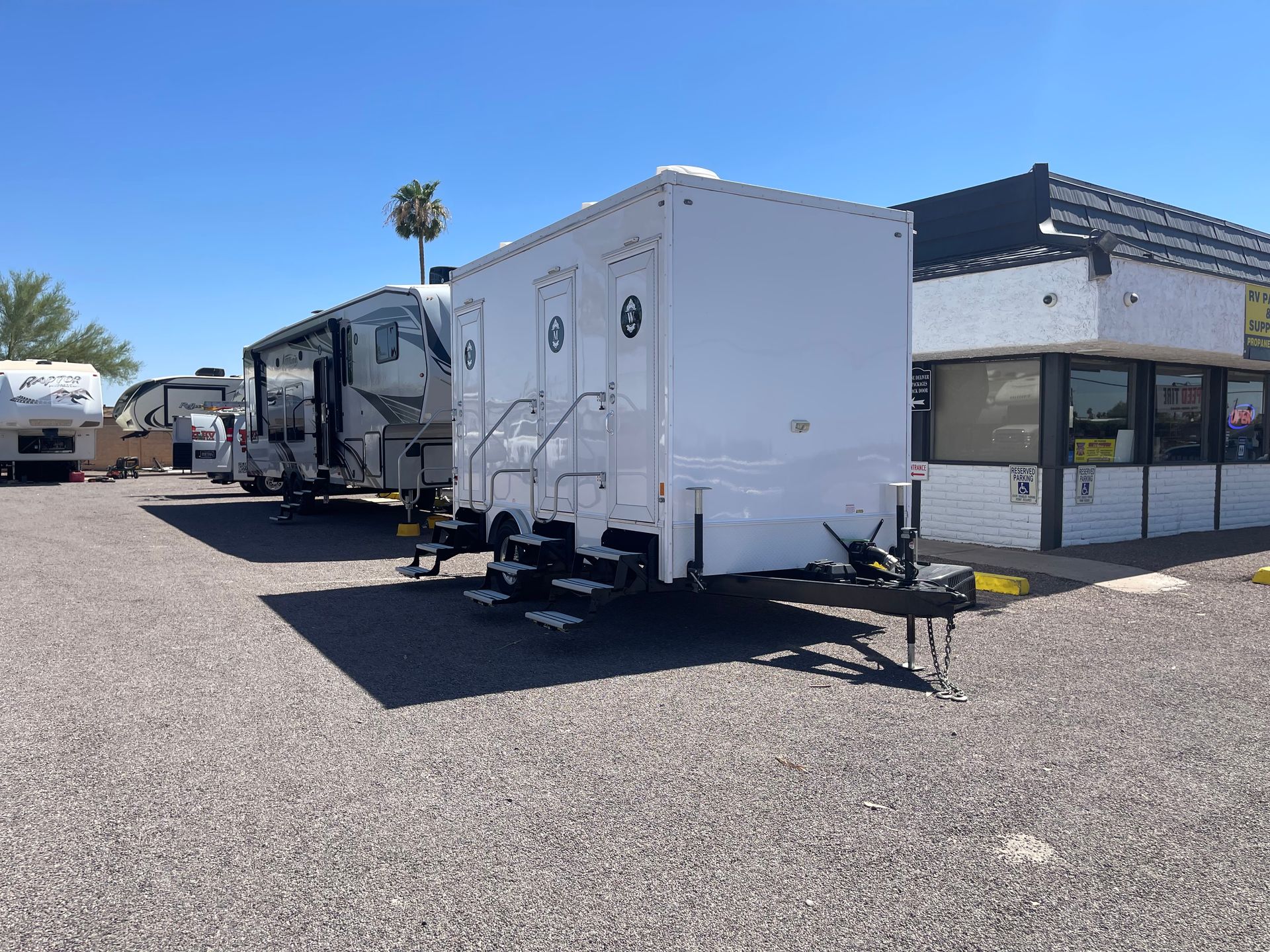 White portable restroom trailer parked on gravel, next to RVs and a building. Blue sky.