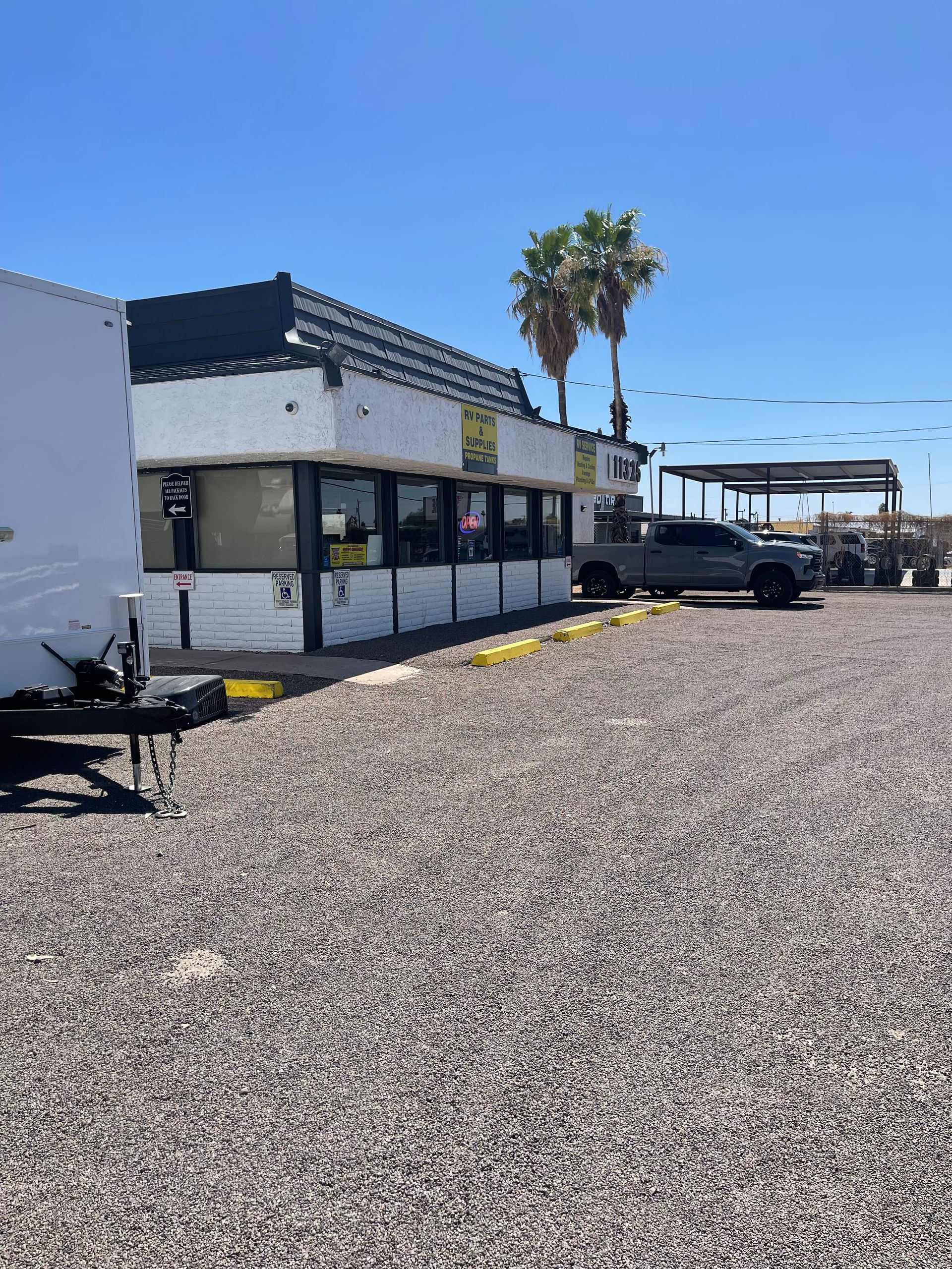 A white building with black trim, parking lot with gray gravel, a truck, and palm trees.