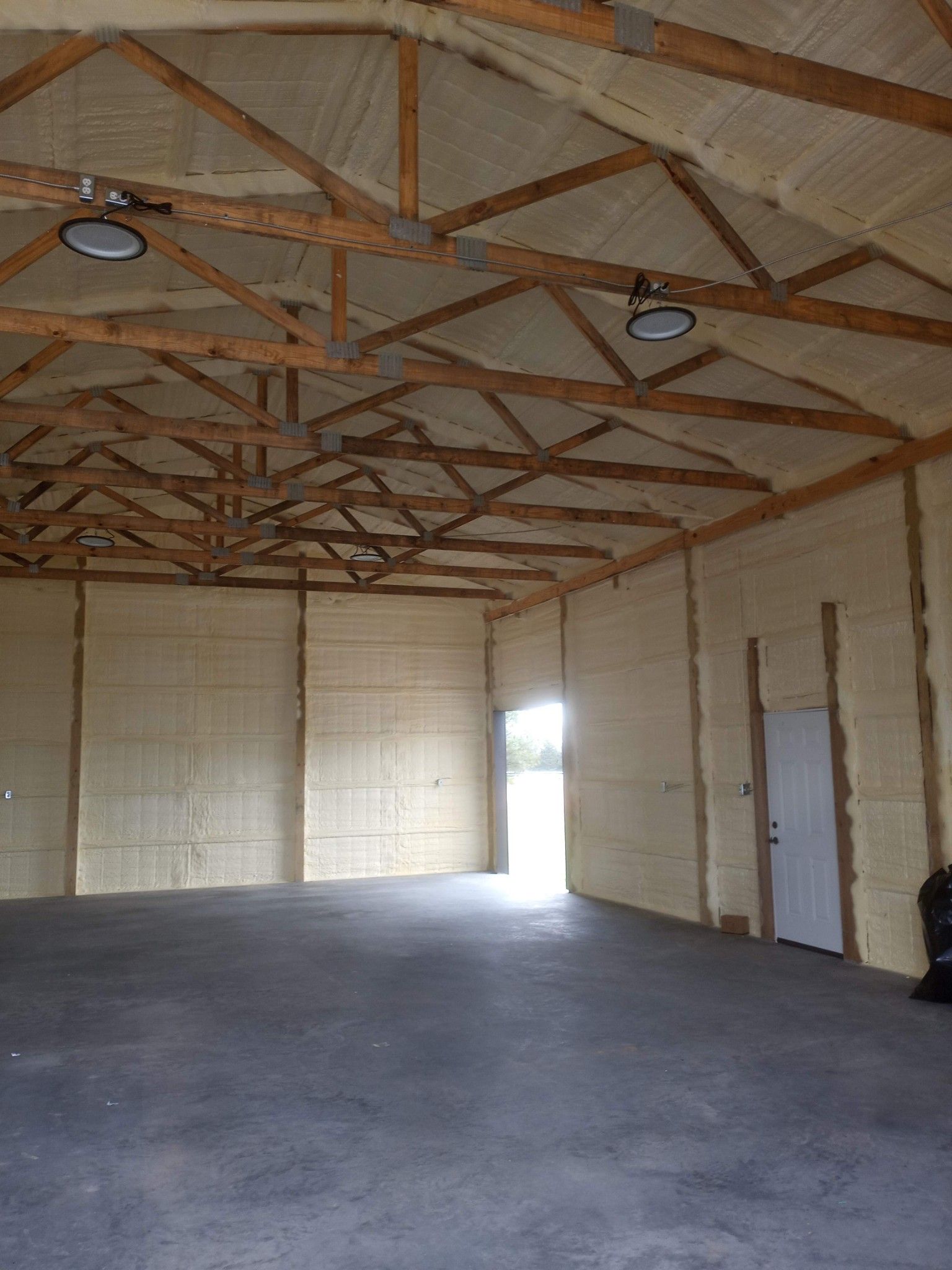 Empty warehouse interior with exposed wooden roof trusses, concrete floor, and a bright doorway at the far end