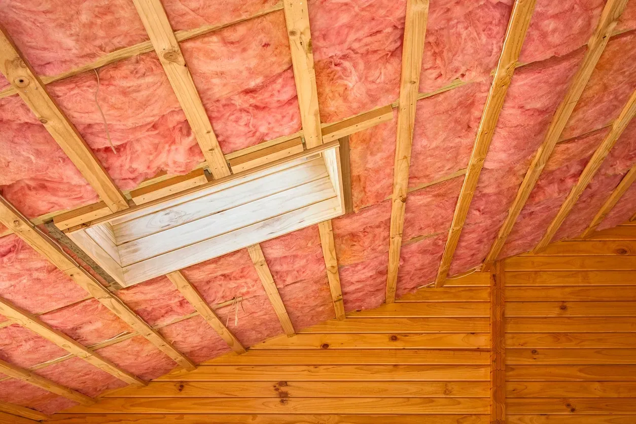 Pink insulation in an unfinished ceiling with wooden beams and a fluorescent light fixture