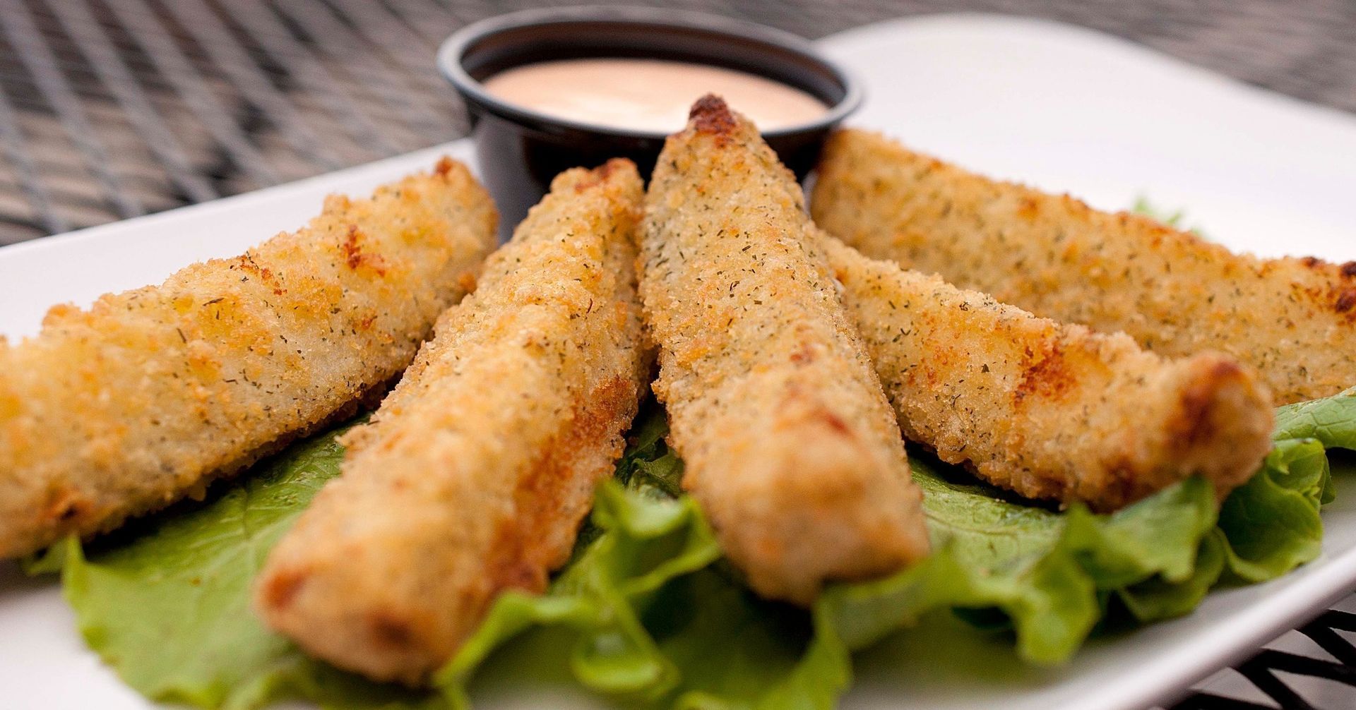 Breaded, fried pickle spears on a bed of green lettuce, served with a side of dipping sauce on a rectangular white plate.