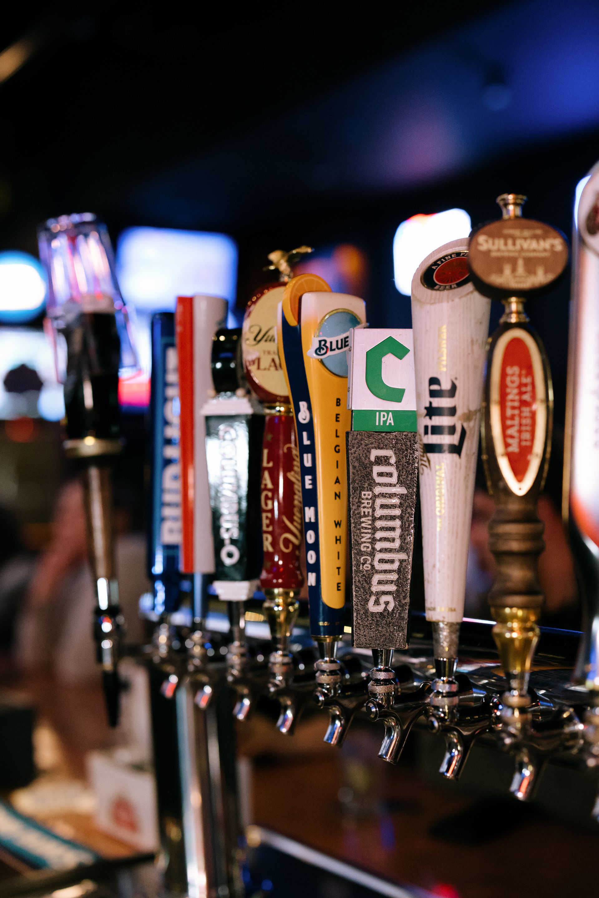 Close-up of beer taps in a bar, various logos visible, stainless steel, dimly lit.