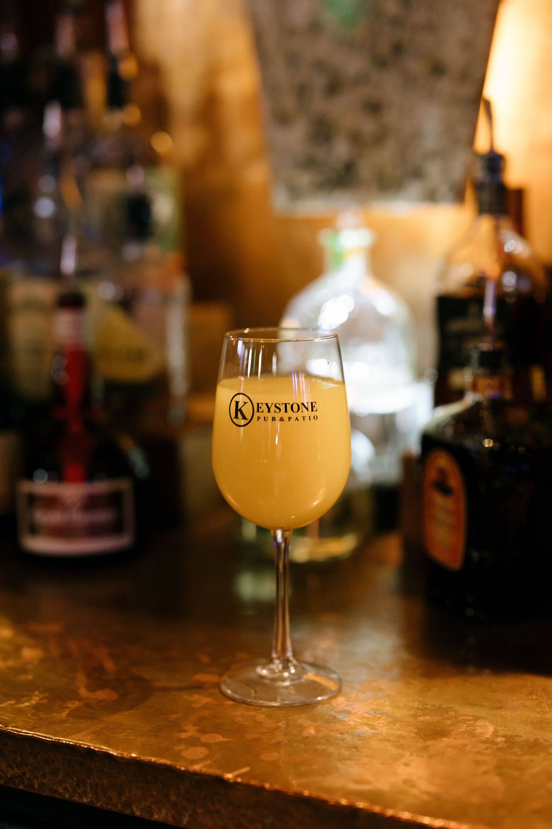 A glass of orange-colored cocktail on a bar, with bottles in the background.
