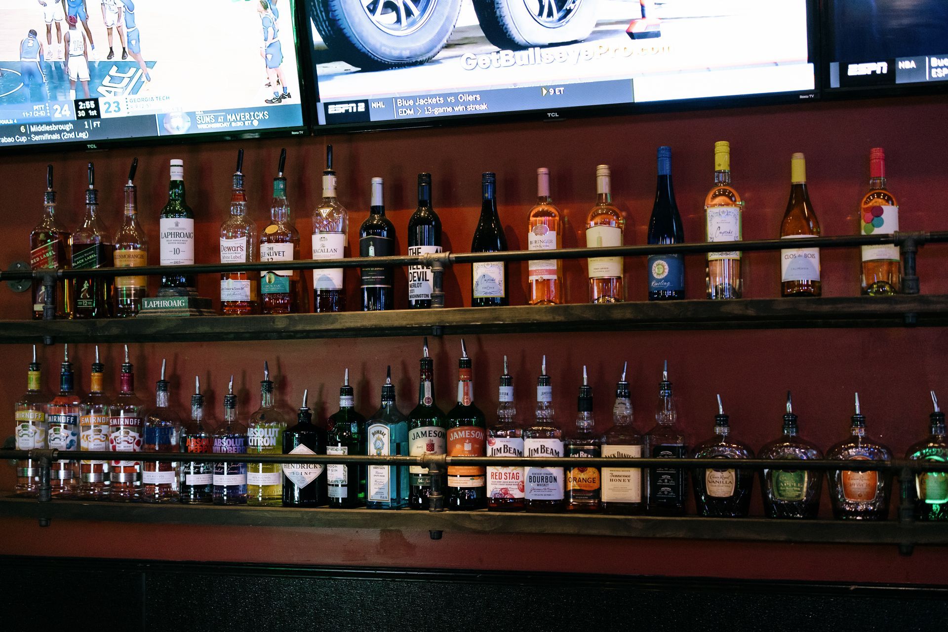 Bar shelves with various liquor bottles, under a TV displaying sports.