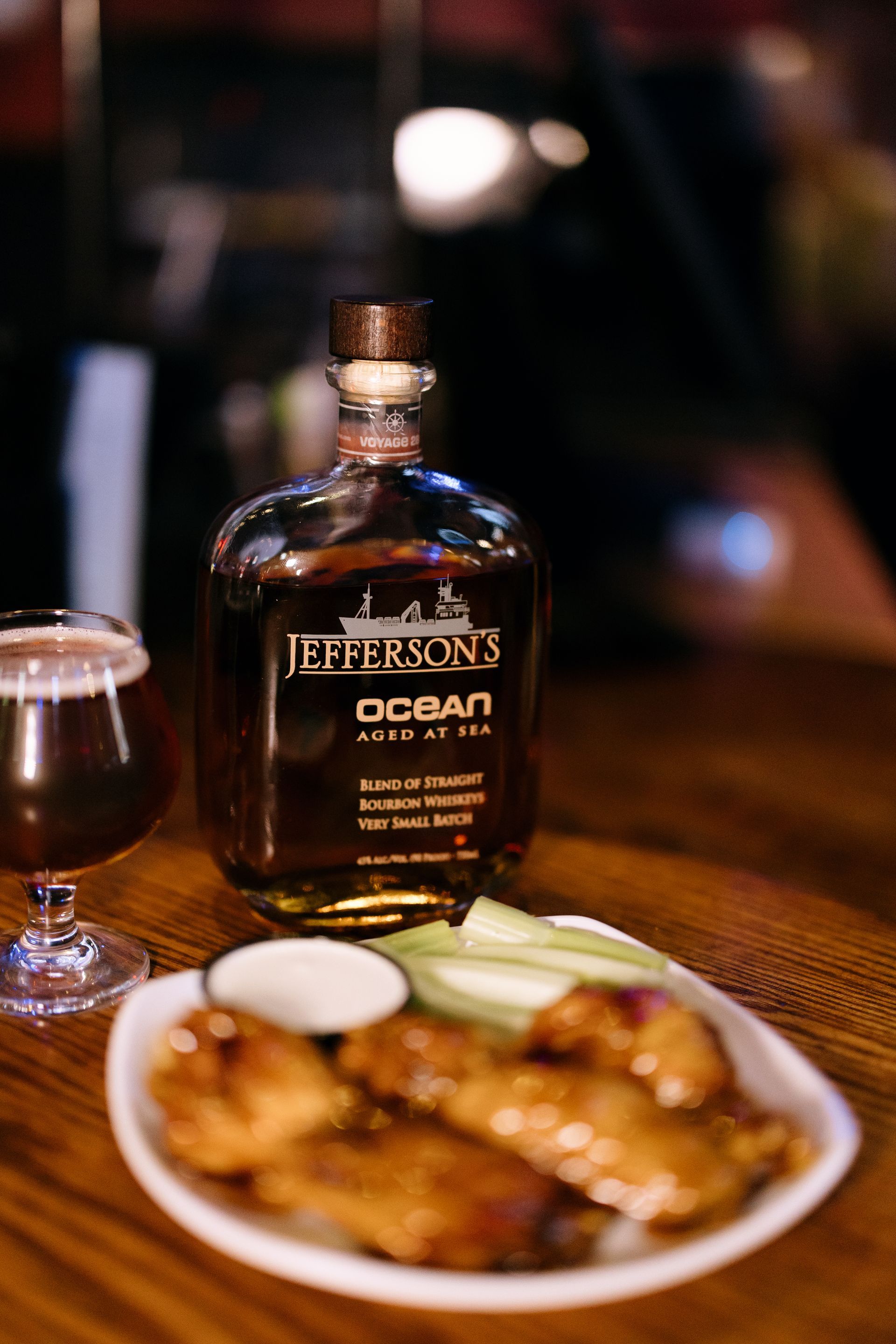 Jefferson's Ocean bourbon bottle with chicken wings, celery, and a beer in a dimly lit bar.