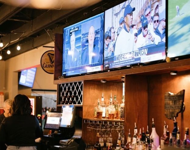 Bar interior with TVs showing news and sports, bartender, liquor bottles.