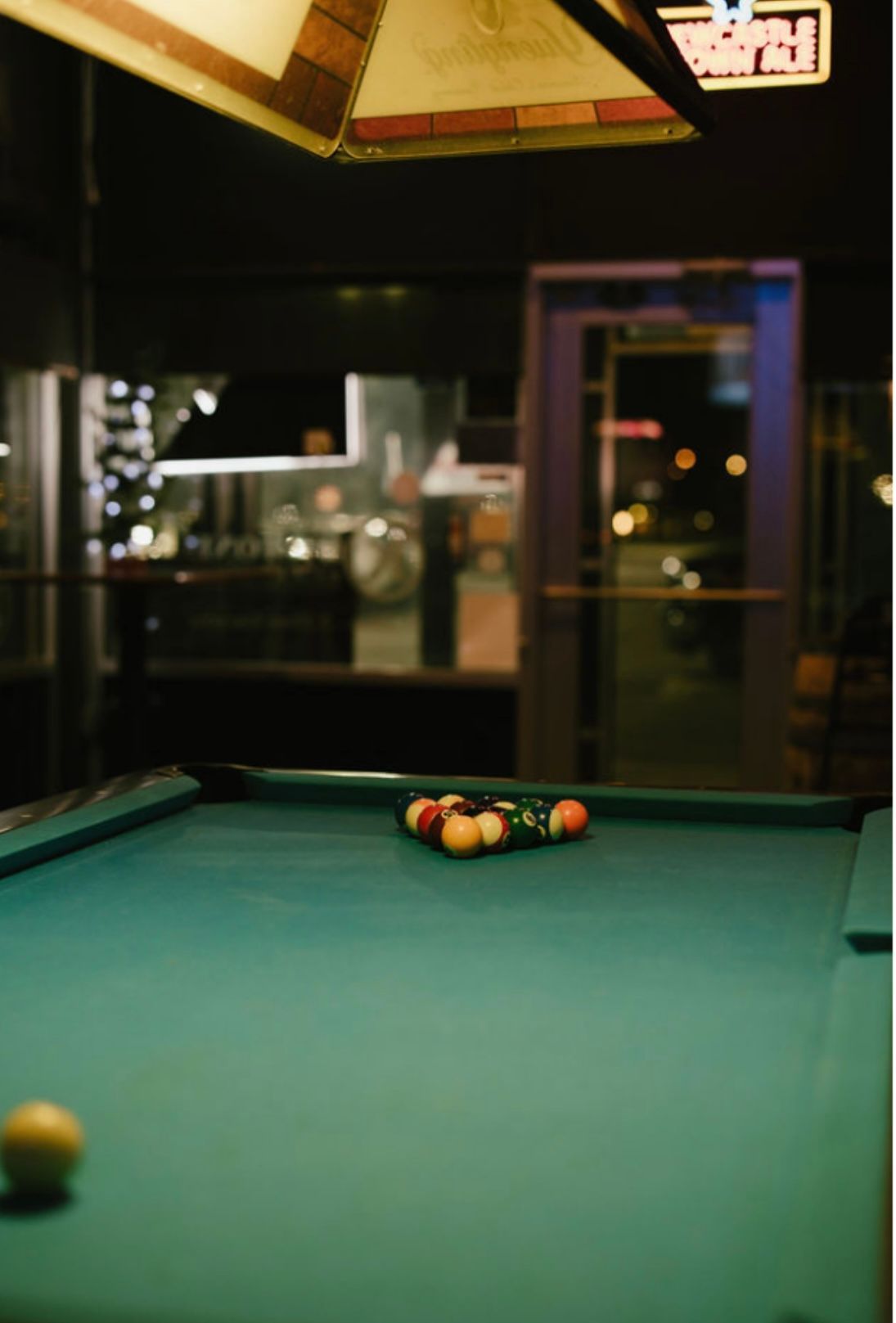 Pool table with racked balls, lit by overhead light, in a dimly lit bar.