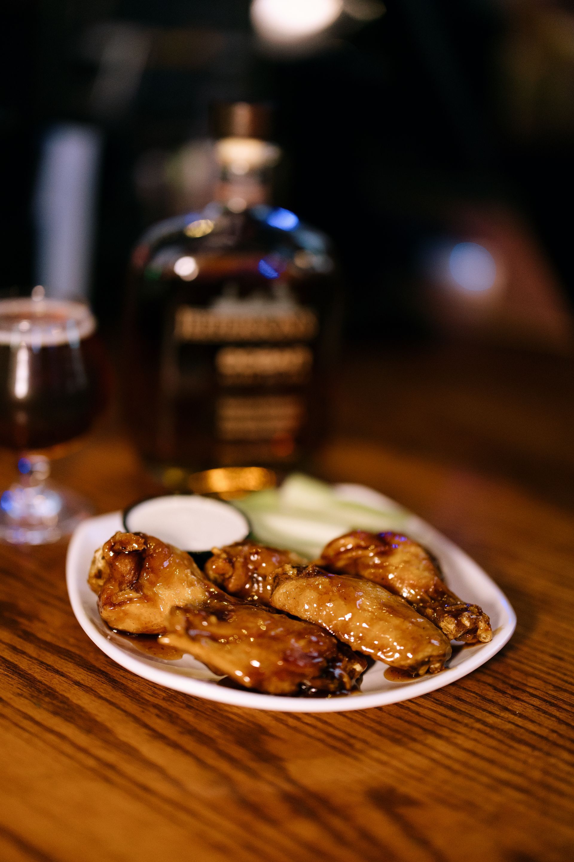 Plate of glazed chicken wings, with celery, dip, and a whiskey bottle on a wooden table.