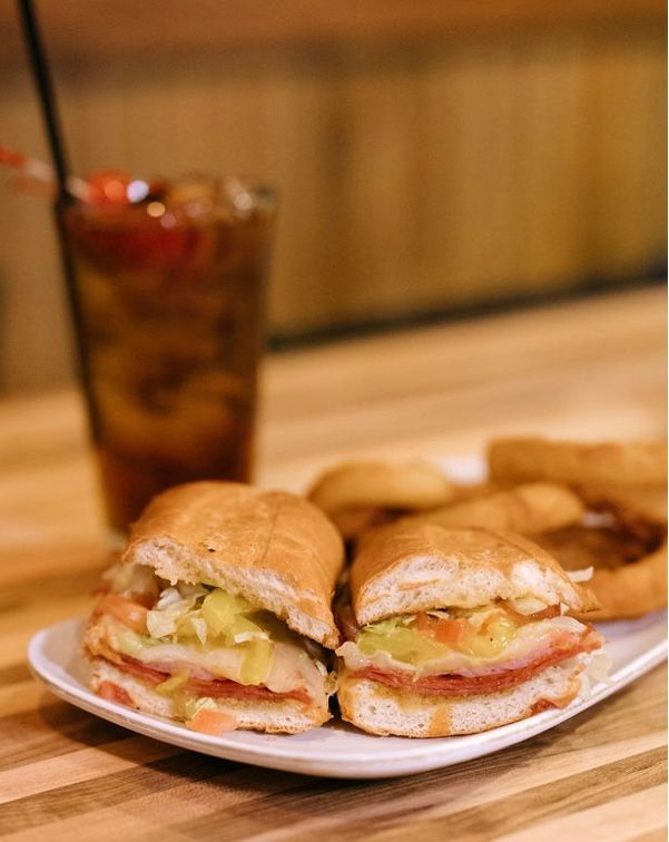 Two submarine sandwiches on a white plate, with a drink in the background, all on a wooden surface.