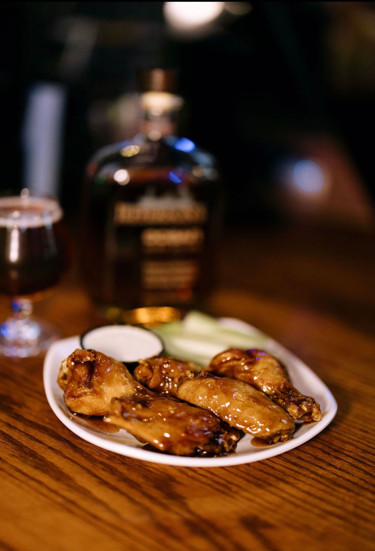 Plate of glazed chicken wings with blue cheese, celery, a whiskey bottle, and a dark beer on a wooden bar.