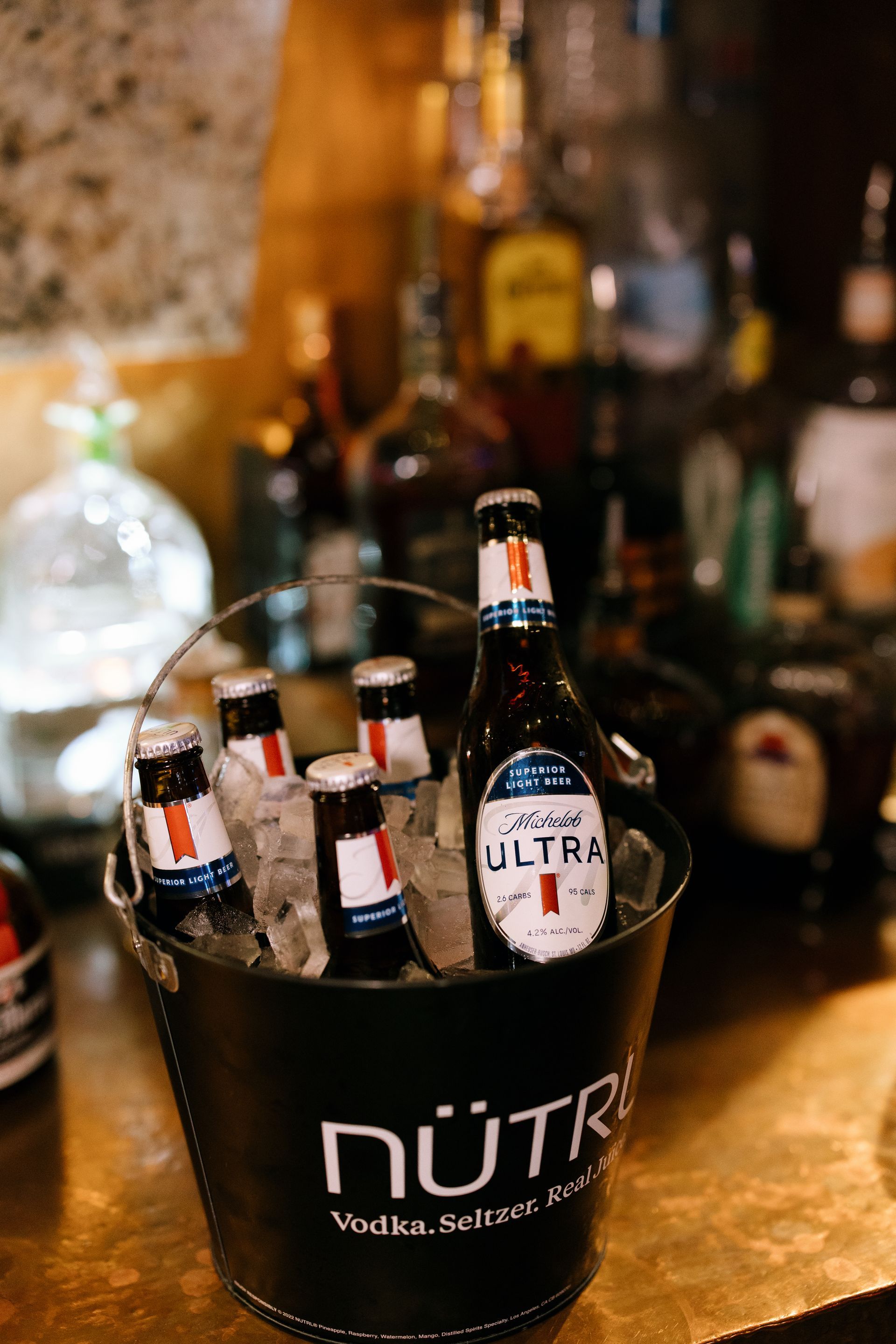 Bucket of beer bottles in ice on a bar, with liquor bottles in background.
