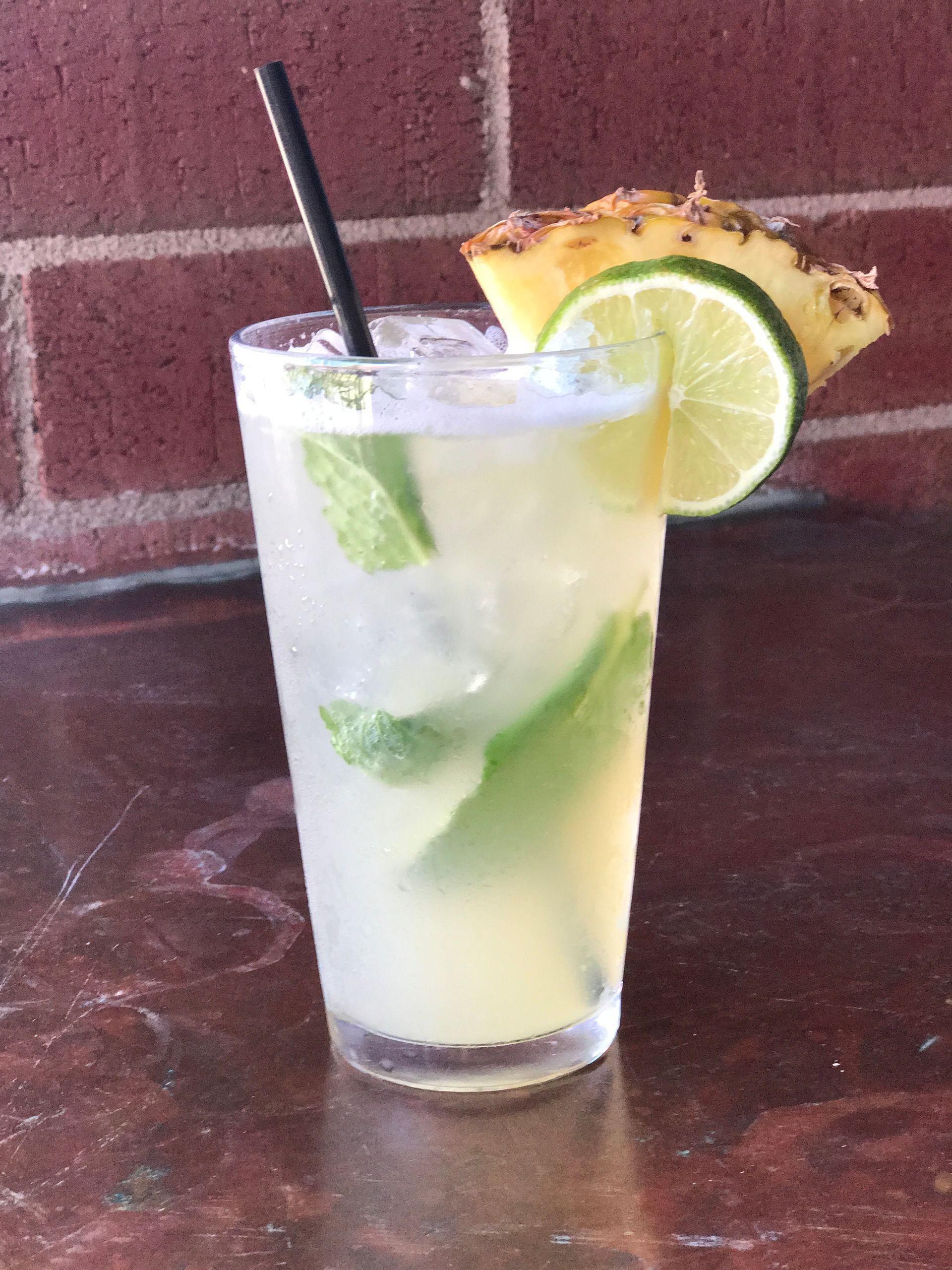 Cocktail with mint and lime garnish, pineapple wedge, black straw, in tall glass on bar.
