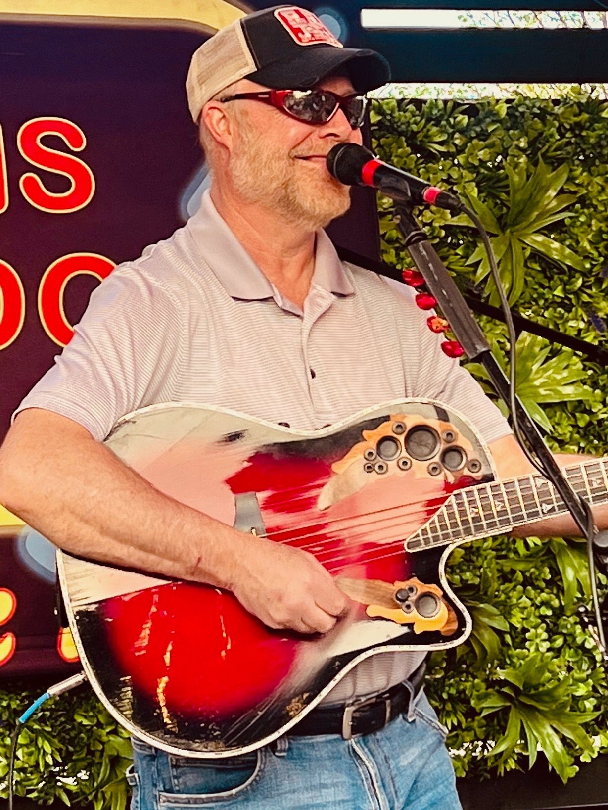 Man playing guitar and singing into a microphone outside, wearing sunglasses and a cap.