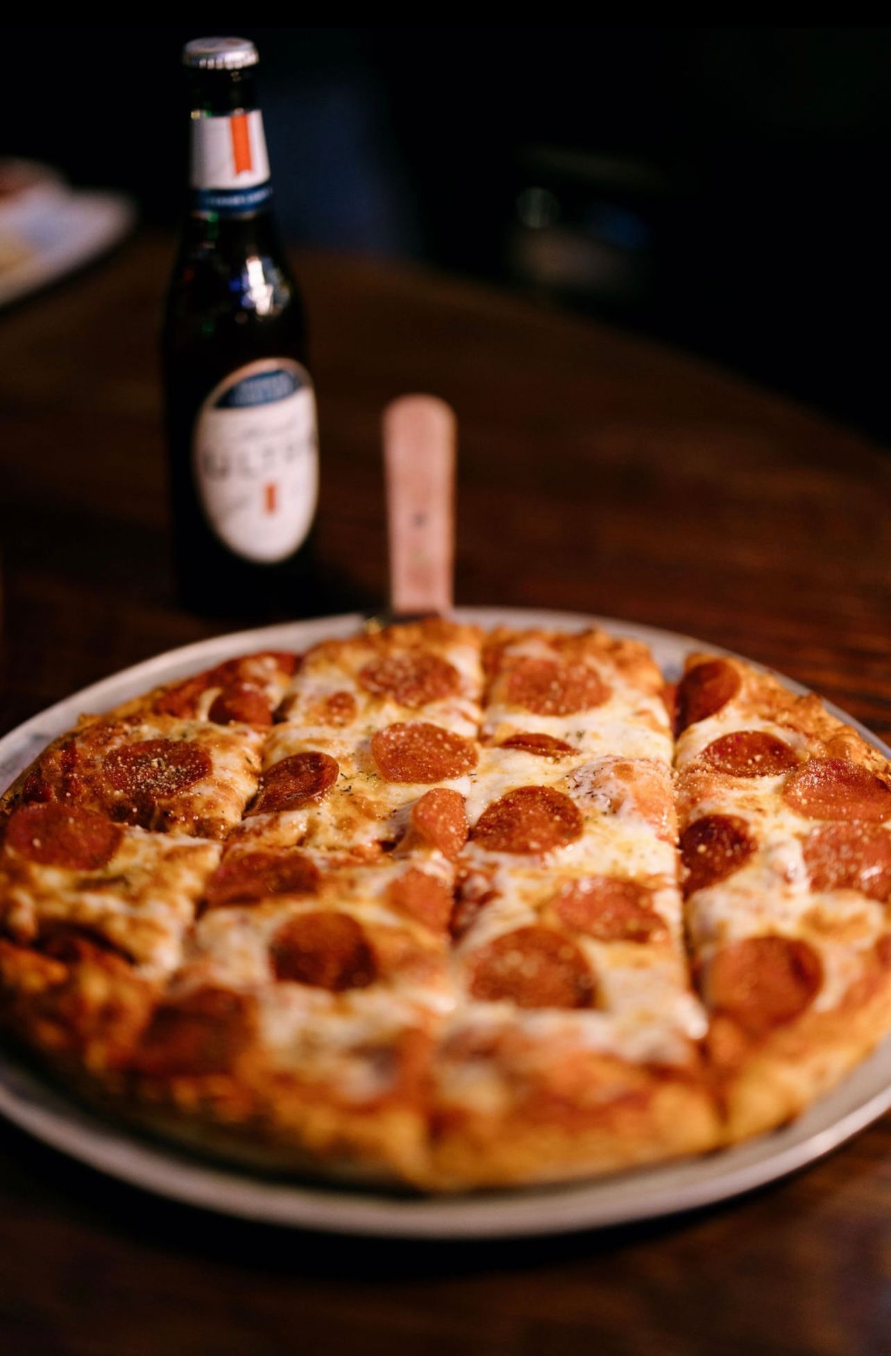 Pepperoni pizza on a plate, a bottle of beer in the background, all on a wooden table.