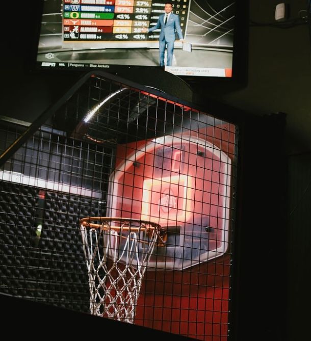 Basketball arcade game with orange illuminated backboard, net hoop, and TV screen in the background.