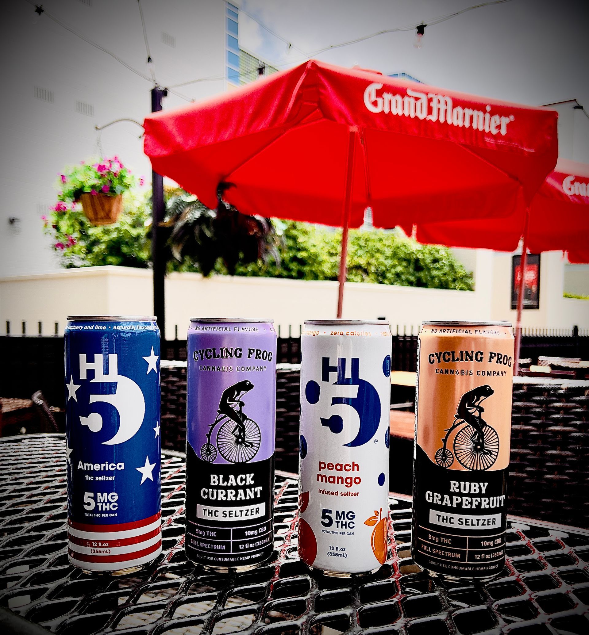 Four Hi5 seltzer cans on a patio table, under a red umbrella.