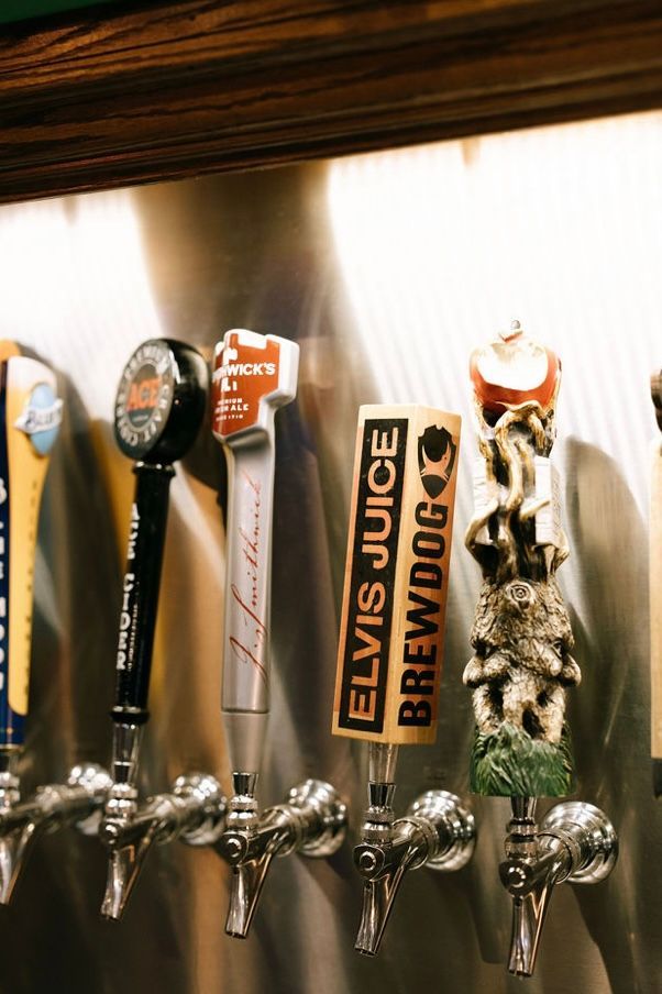 Beer taps with colorful logos on a stainless steel wall in a bar.