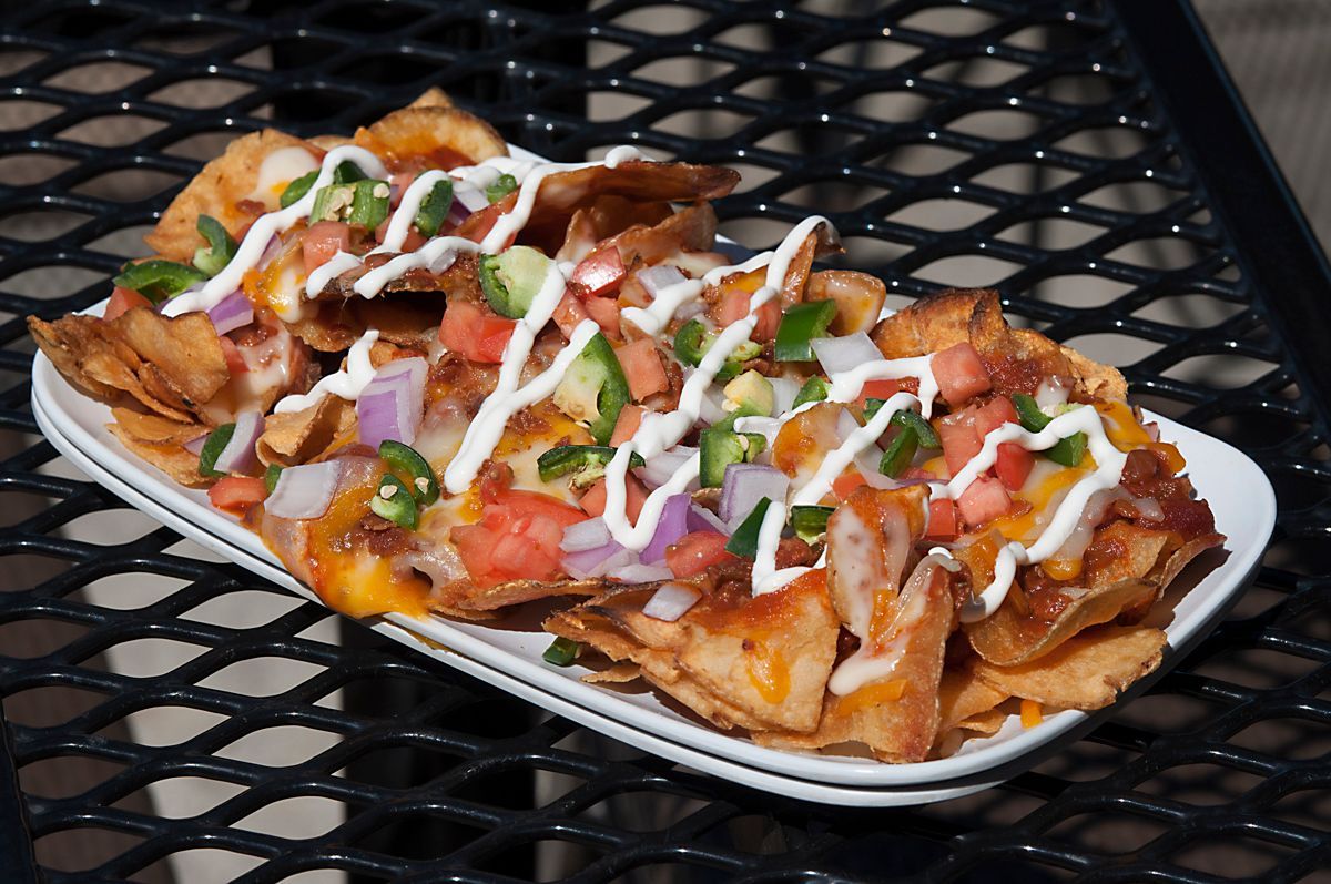 Loaded potato chip nachos, topped with cheese, tomatoes, jalapeños, onions, and a white sauce on a white plate.