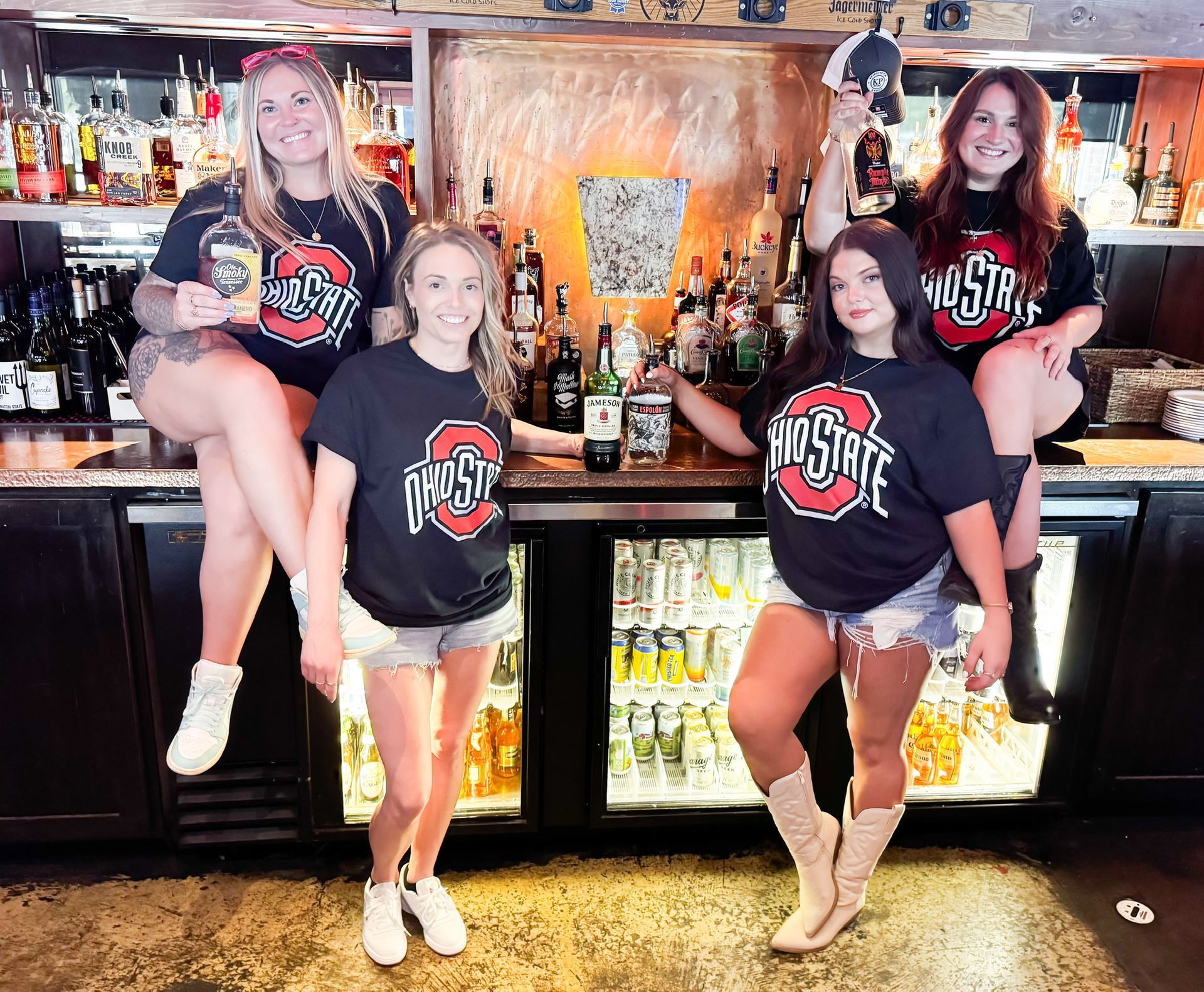 Four people wearing Ohio State t-shirts pose behind a bar with alcohol bottles. 