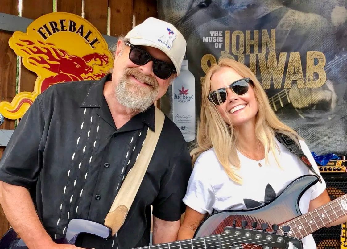 A man and a woman smile, holding guitars. They stand outside near a sign. 