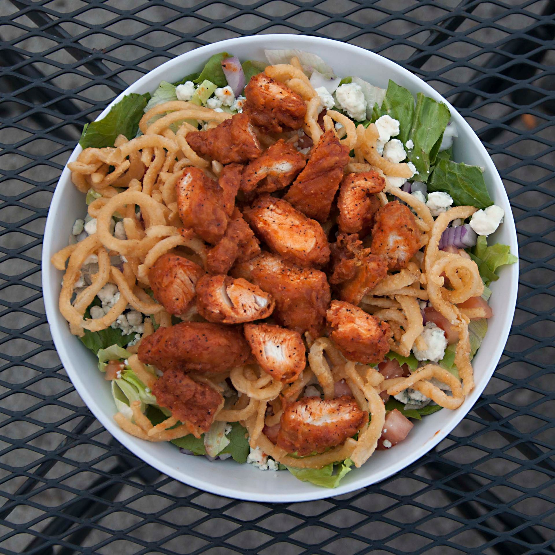 Salad bowl with fried chicken, onion straws, lettuce, and feta cheese on a patio table.