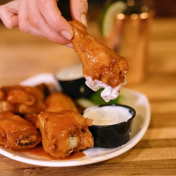 A person dipping a glazed chicken wing into a small cup of white dip, beside more wings.