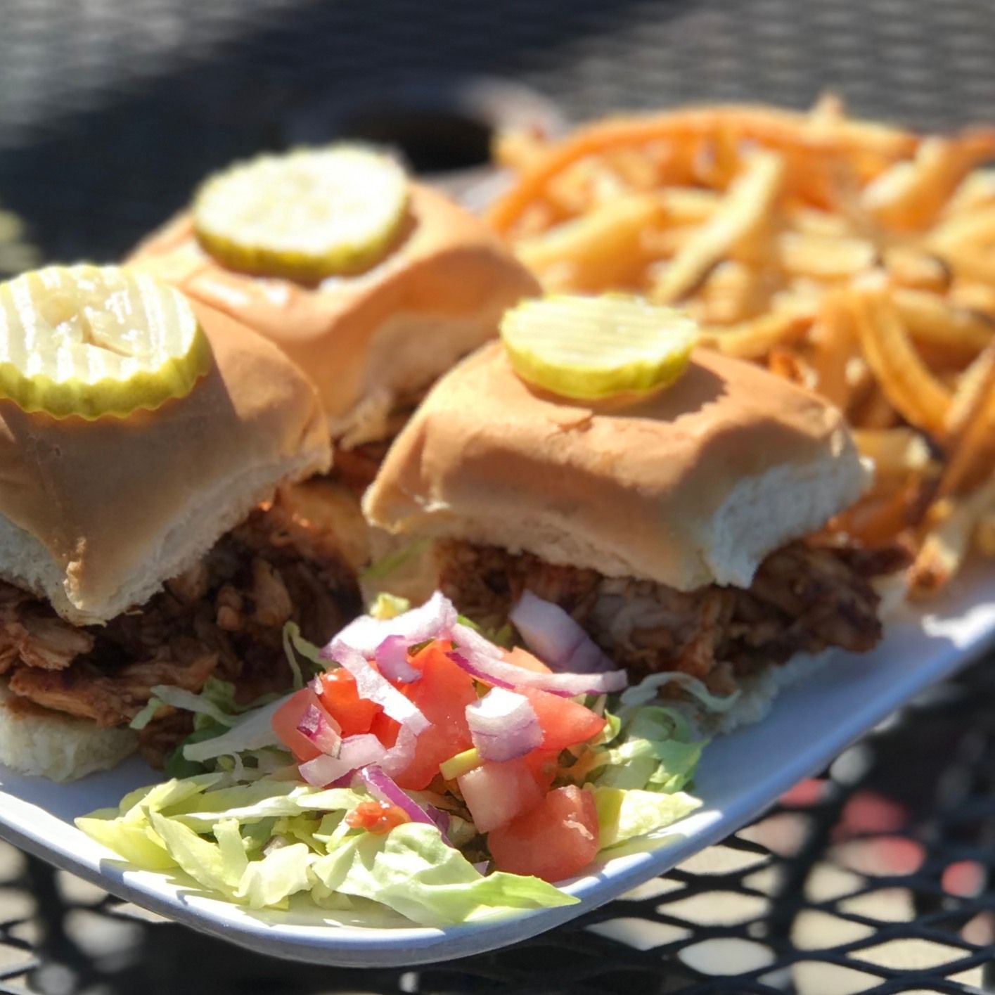 Three sliders topped with pickles, side of fries, and diced tomatoes on a white plate.