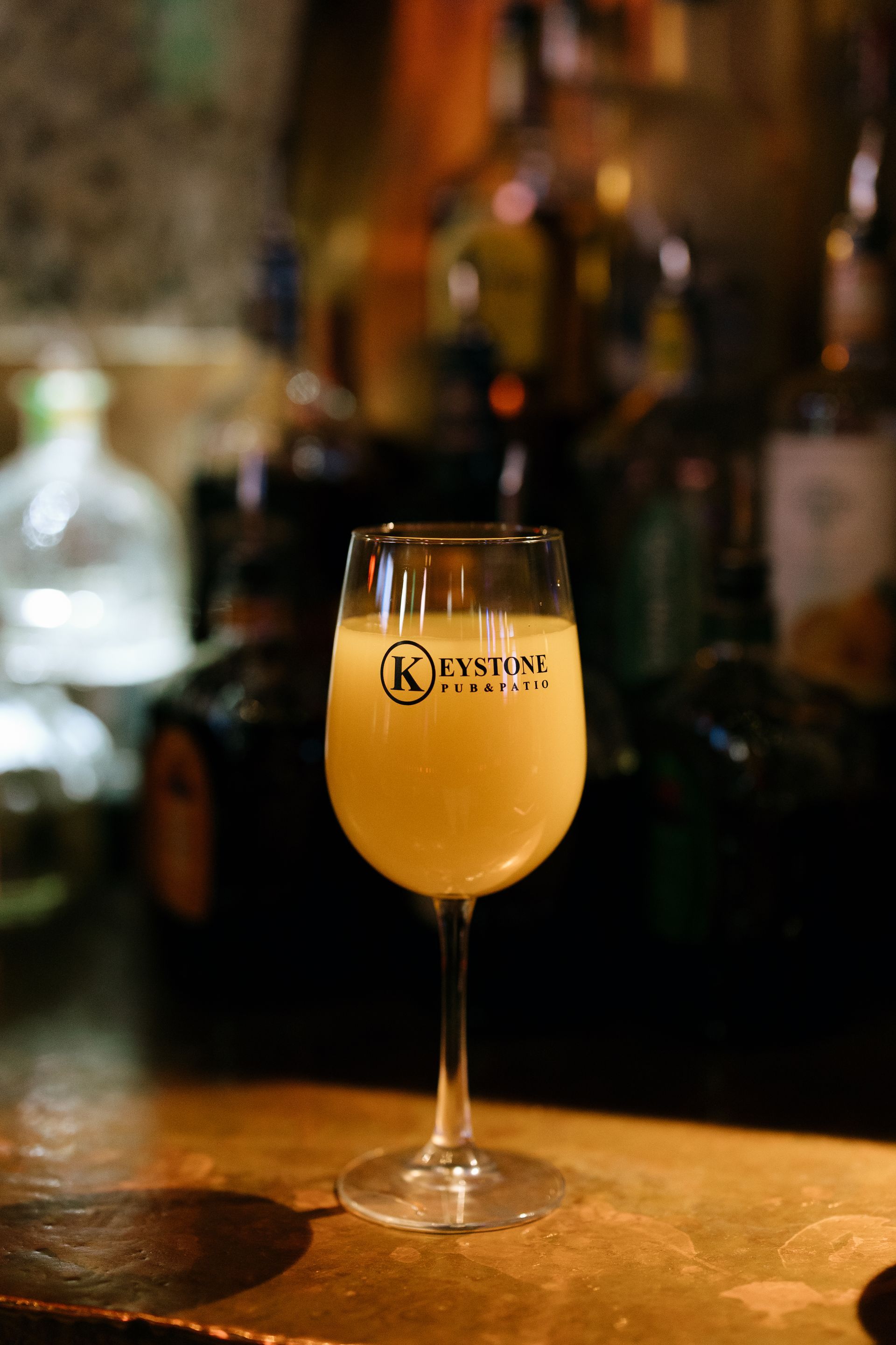 A stemmed glass of yellow cocktail sits on a bar, blurry liquor bottles in the background.