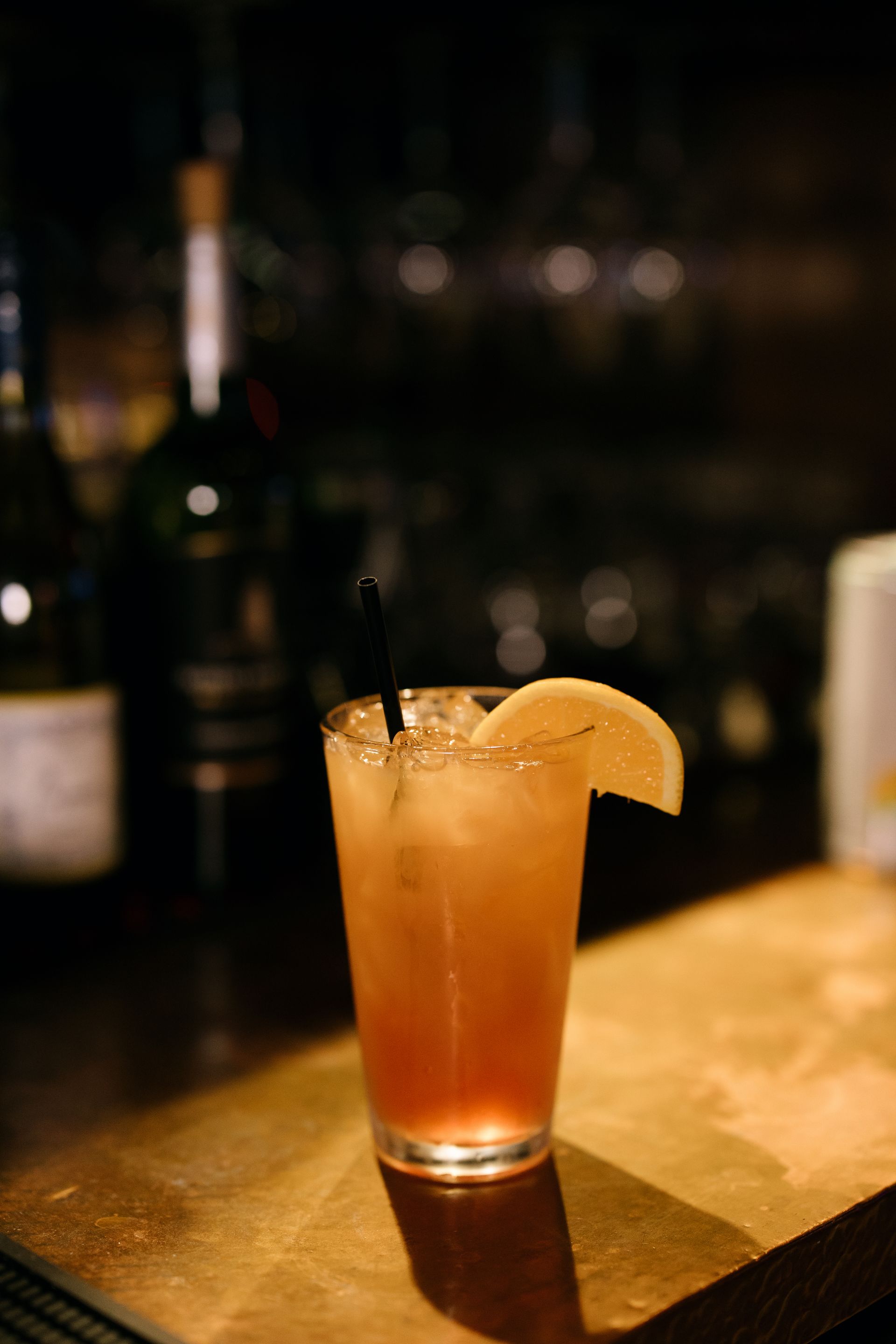 Cocktail in a tall glass with ice and lemon slice, on a bar with bottles in background.