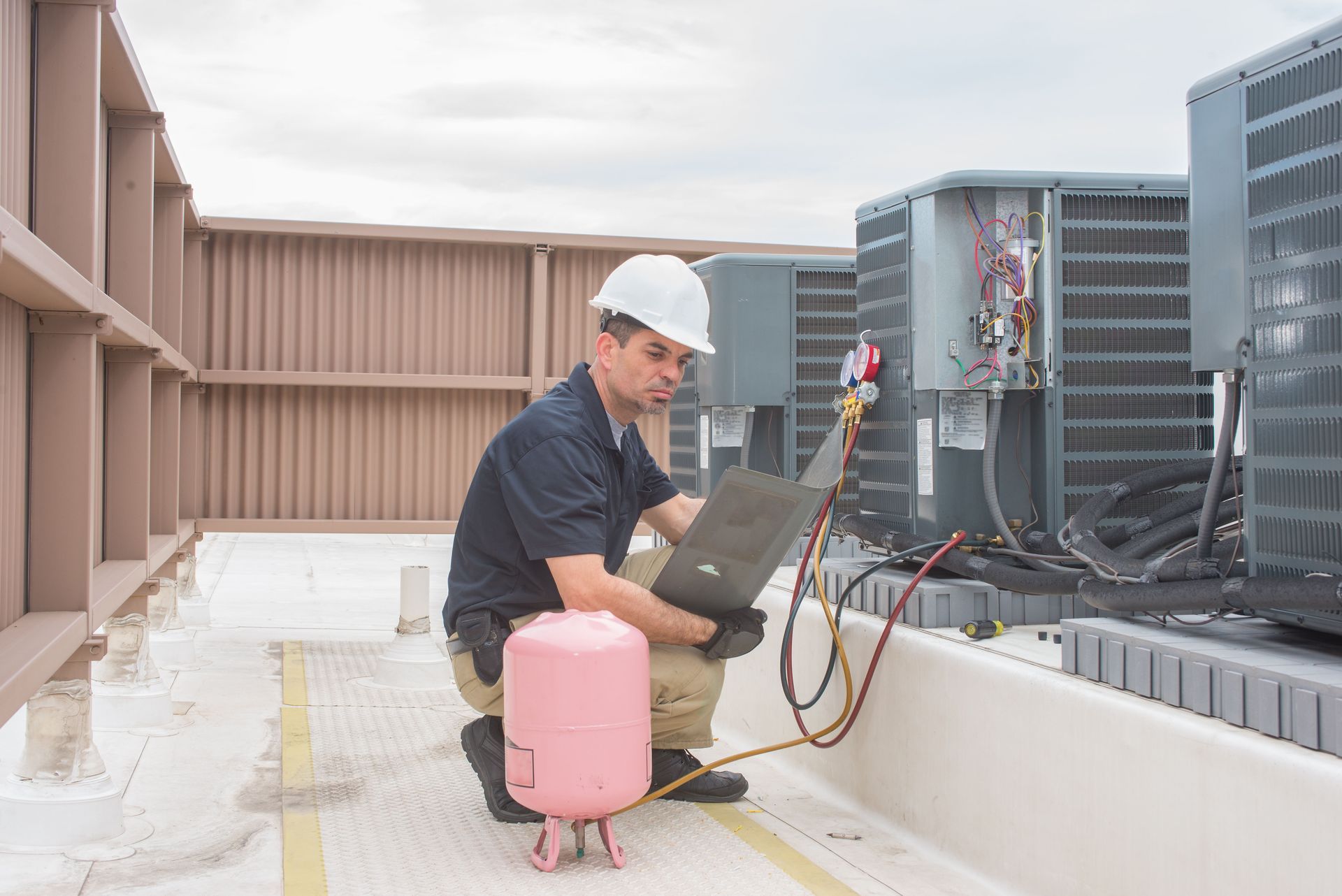 A technician in a hard hat kneeling on a rooftop, using a laptop to service industrial HVAC units with a refrigerant tank.
