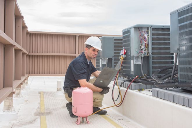 A technician in a hard hat kneeling on a rooftop, using a laptop to service industrial HVAC units with a refrigerant tank.