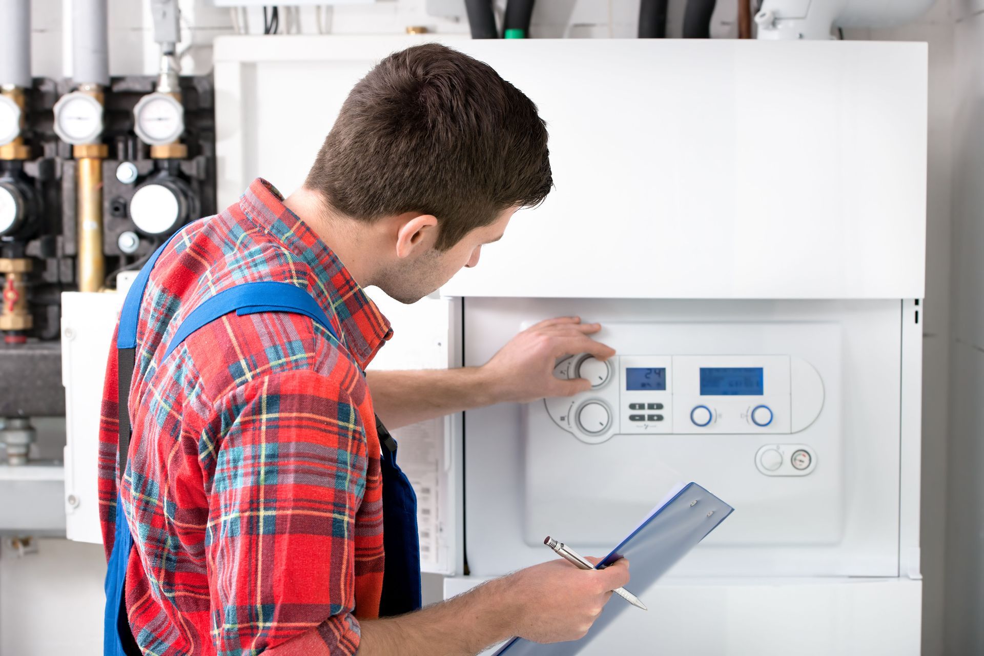 A technician in a red plaid shirt and blue overalls adjusting the settings on a white industrial boiler with a clipboard.