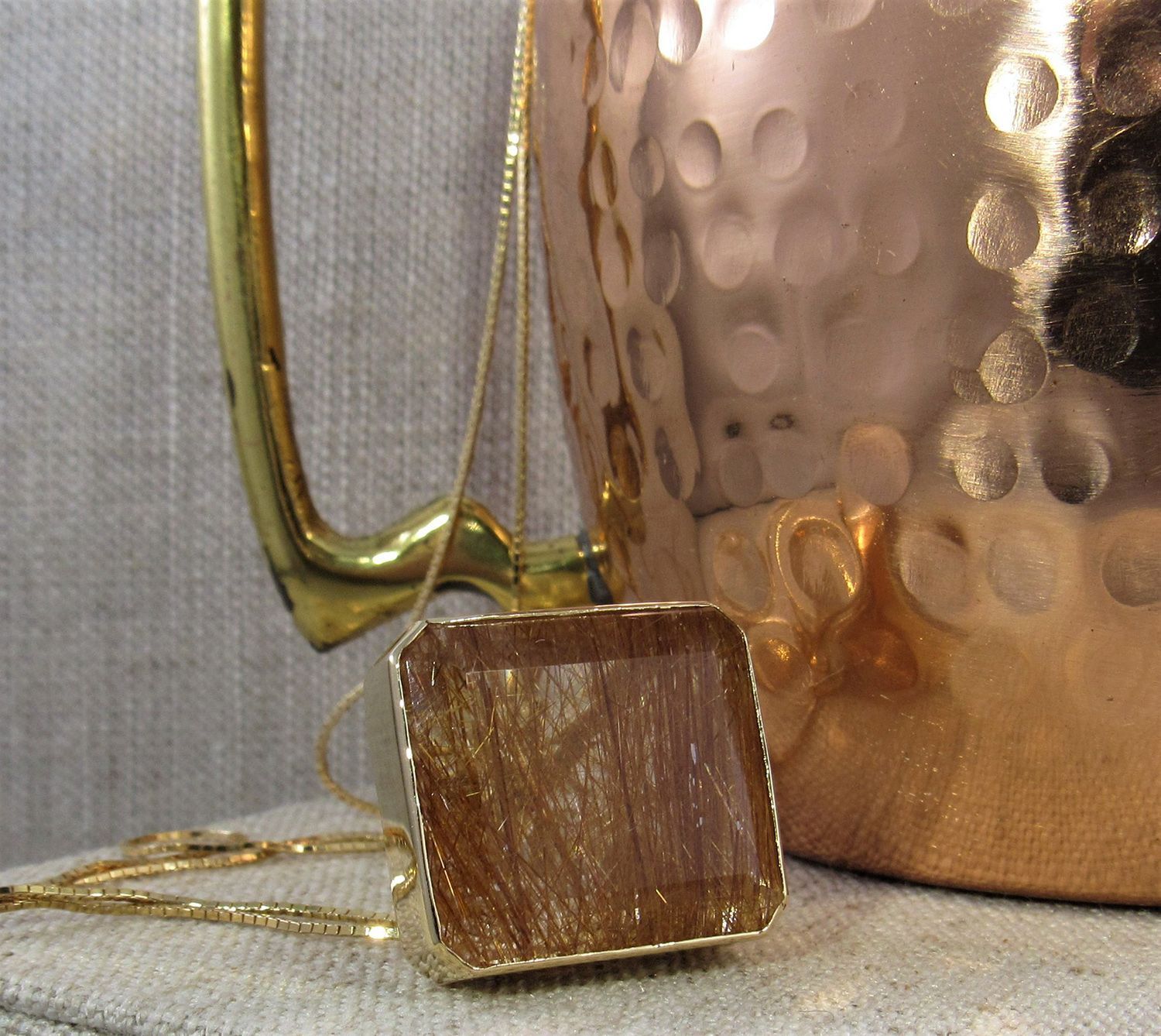 A gold necklace with a square pendant is next to a copper pot