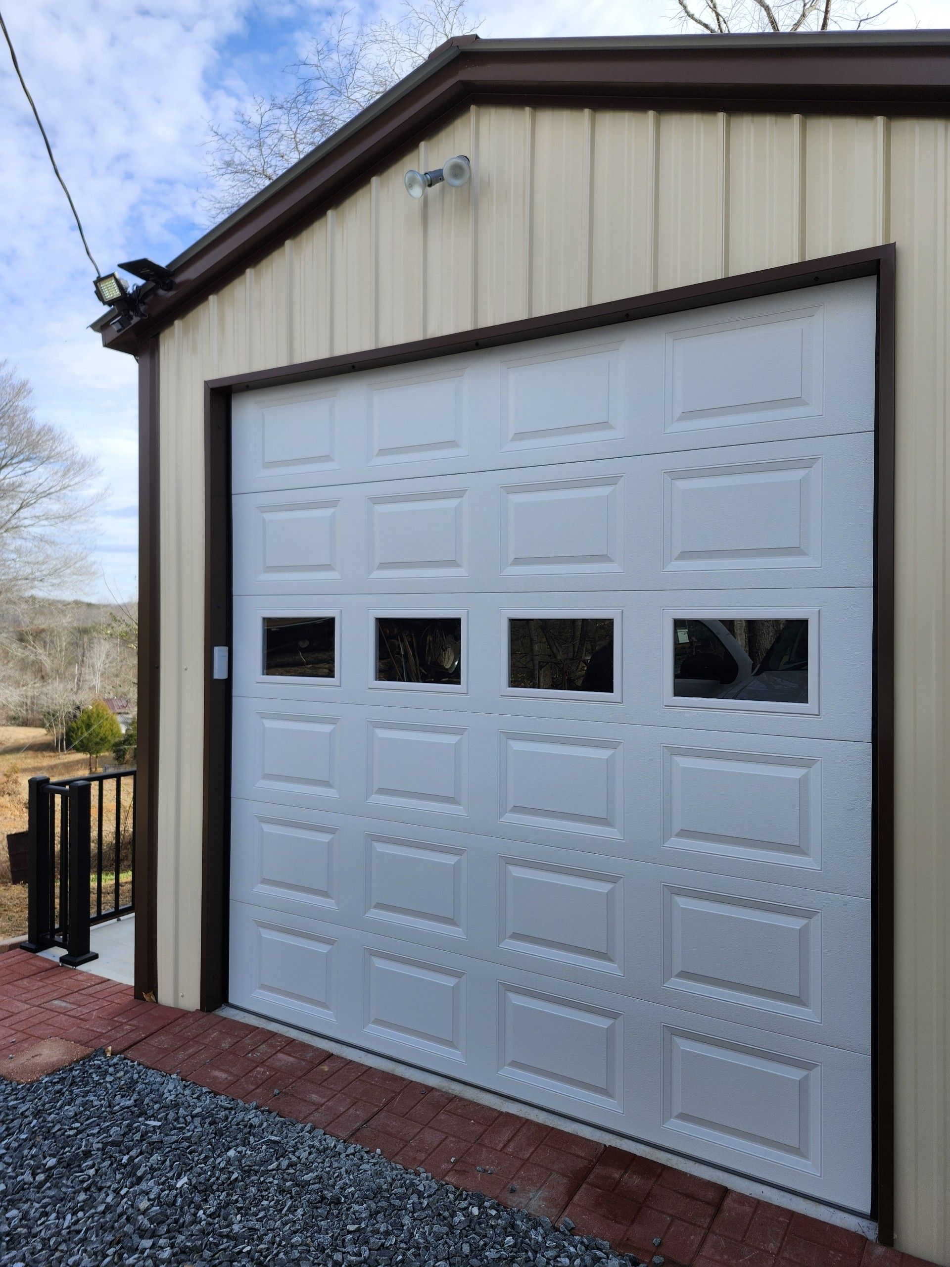 White garage door on a tan metal building with brown trim and windows. Brick and gravel ground outside.