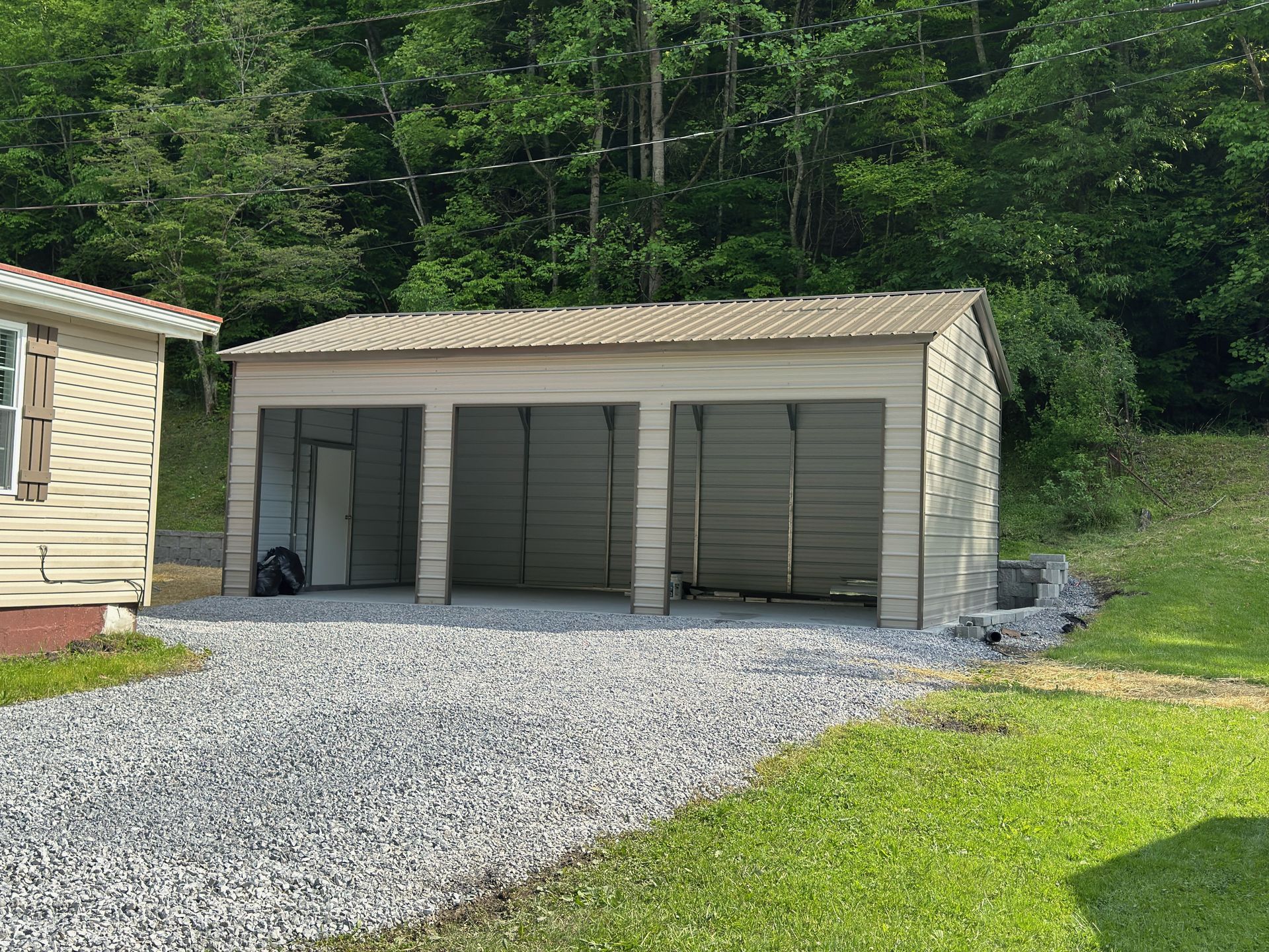Three-bay garage with gray siding and metal roof on a gravel driveway, next to a house and grassy area.
