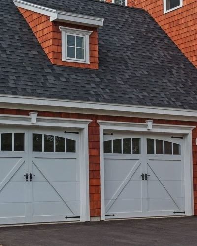Two white garage doors with black handles, a red shingled facade, and a dormer.
