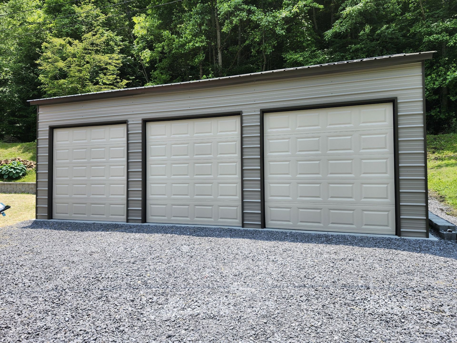 Three-bay gray metal garage with tan doors and a sloped roof, gravel driveway, and trees in the background.