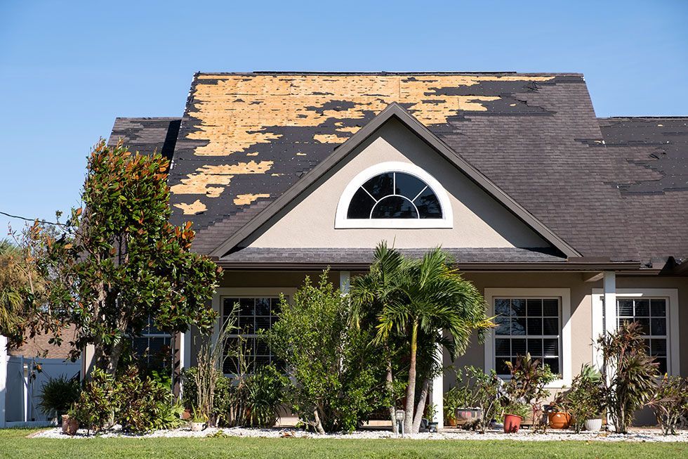 A single-story house with significant wind damage, showing large sections of shingles missing from the roof.