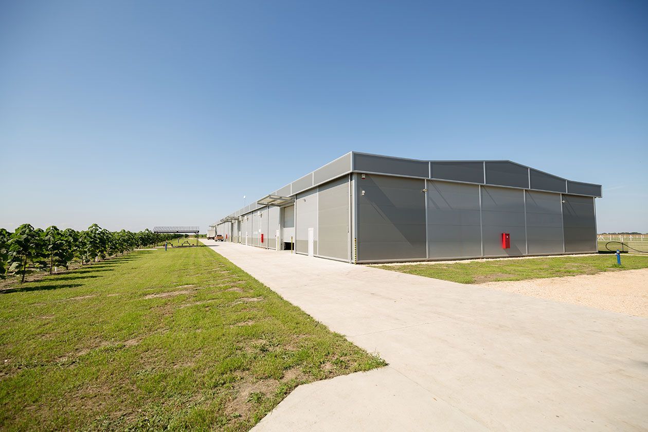 A long, gray industrial metal building stands on a concrete driveway next to a grassy field with young trees.
