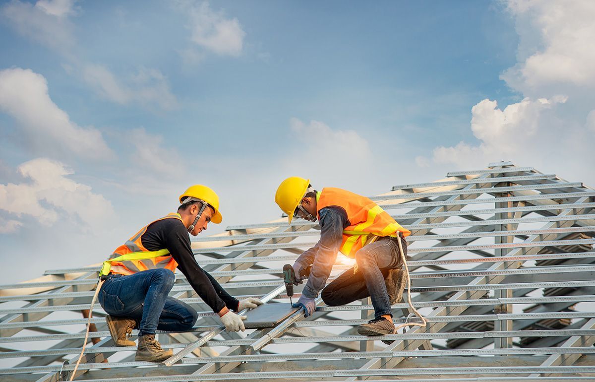 Two construction workers in yellow hard hats and high-visibility vests install roof tiles on a metal frame.