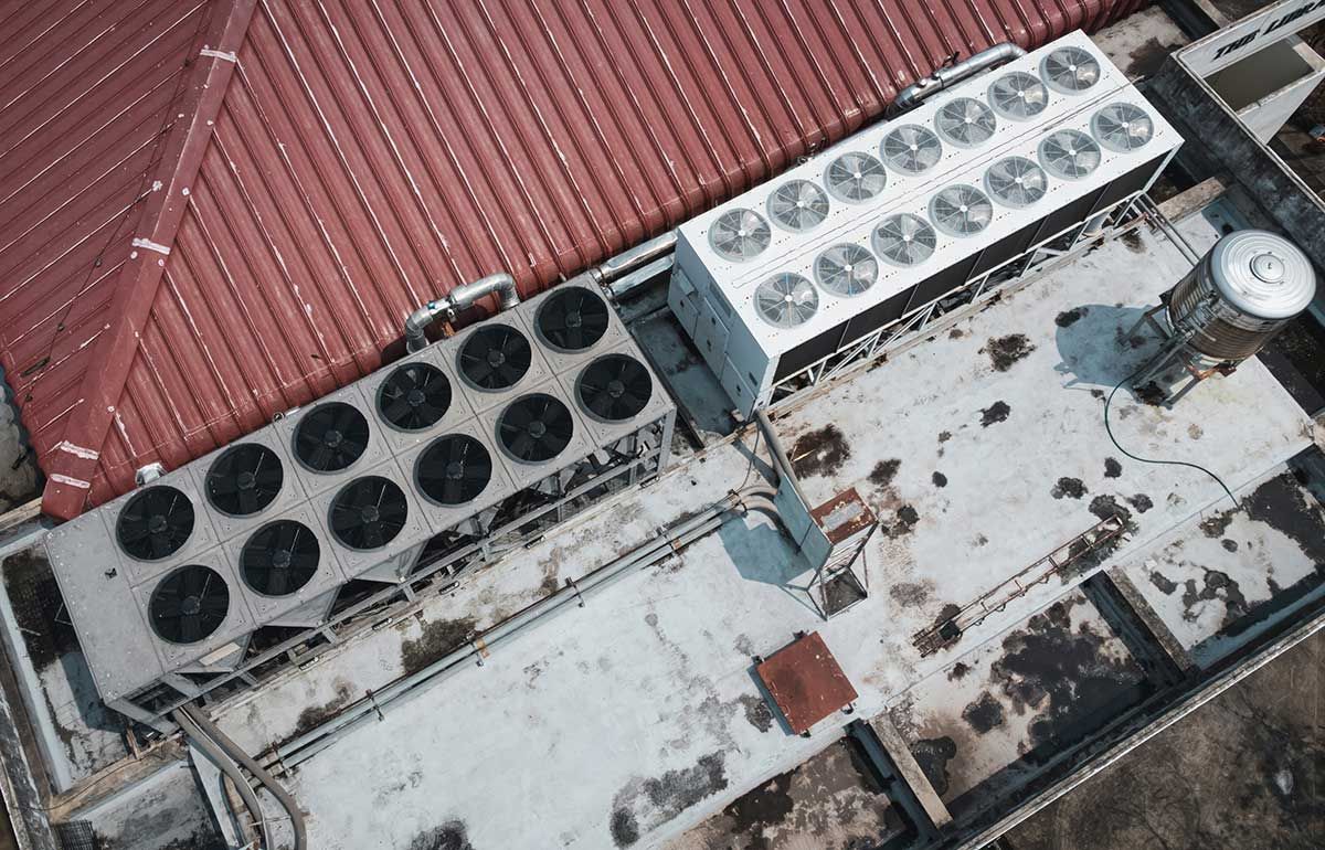Aerial view of two large industrial HVAC cooling units on a flat, weathered gray roof next to a sloped red tiled roof.