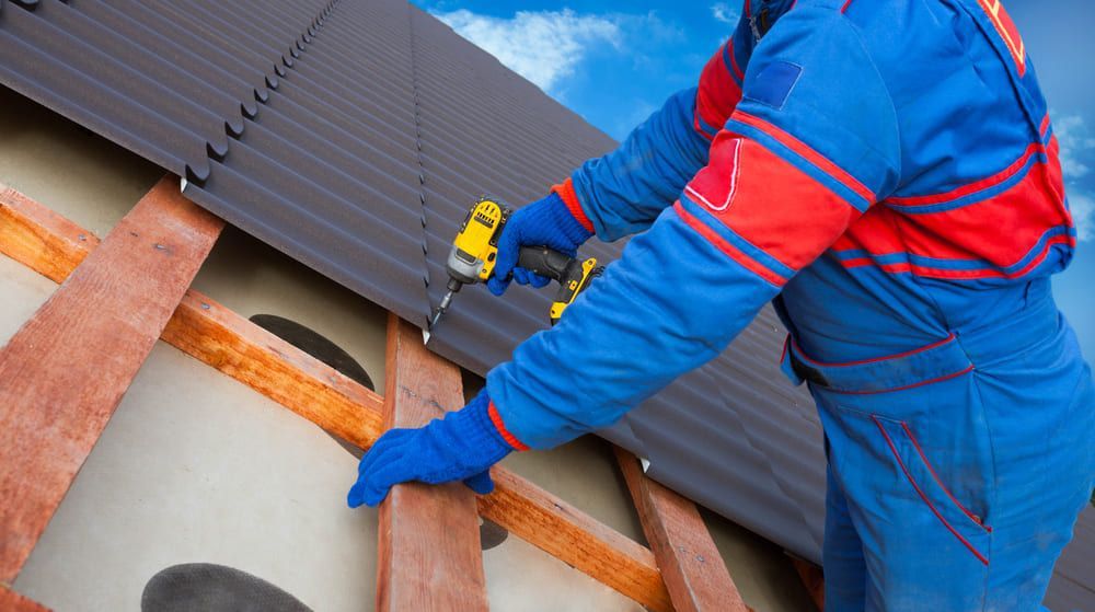 A construction worker in a blue uniform and gloves uses a power drill to secure roofing panels to wooden beams.