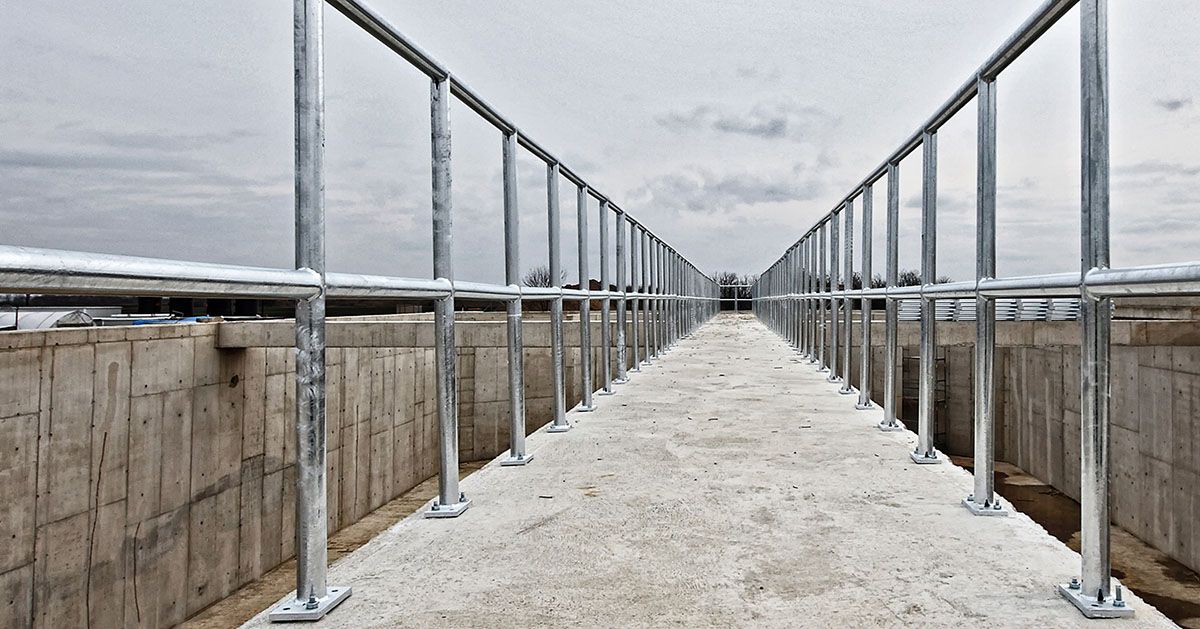 A narrow concrete walkway lined with galvanized steel railings, centered between two concrete walls under a cloudy sky.