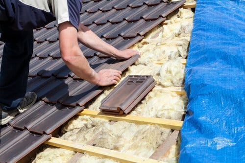 A person installing brown roof tiles over yellow insulation on a sloped roof next to a blue tarp.
