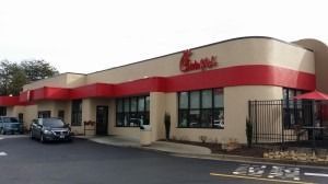 A tan and red Chick-fil-A restaurant exterior with a parking lot in the foreground under a cloudy sky.