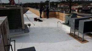 Two workers apply a white roof coating on a commercial rooftop with HVAC units in a suburban area.