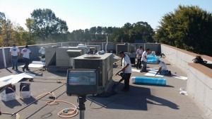 A group of workers in white shirts install HVAC units on a flat building rooftop on a sunny day.