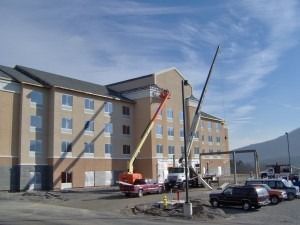 A construction crew uses a yellow boom lift and a crane to work on the exterior of a tan, multi-story hotel building.
