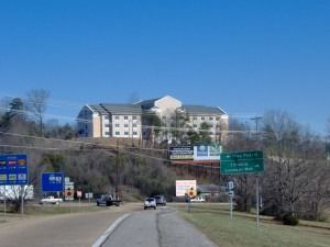 A multistory hotel sits atop a grassy hill overlooking a road with highway signs and moving vehicles under a clear sky.