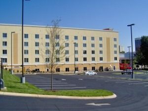 A five-story beige hotel building behind a large, mostly empty asphalt parking lot under a clear blue sky.